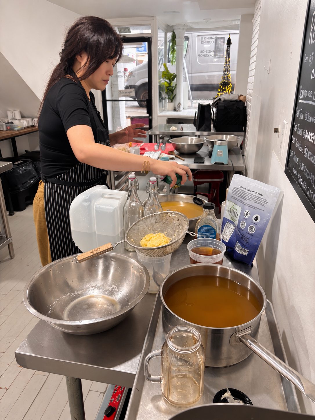 A woman in a striped apron prepares a batch at a steel kitchen counter with swing-top bottles, a strainer and a pot of amber liquid.