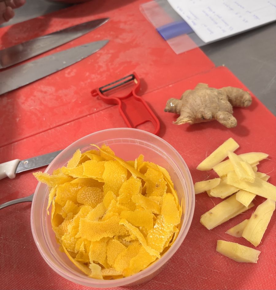 A container of fresh citrus peel beside whole and sliced ginger, a red peeler and knives on a red cutting board.
