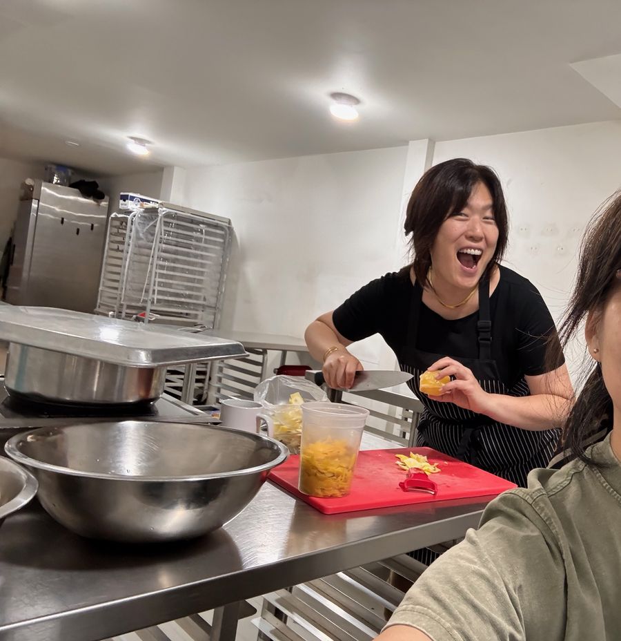 A woman in an apron laughs while cutting citrus at a steel prep table in a commercial kitchen, a tub of peel on a red cutting board.
