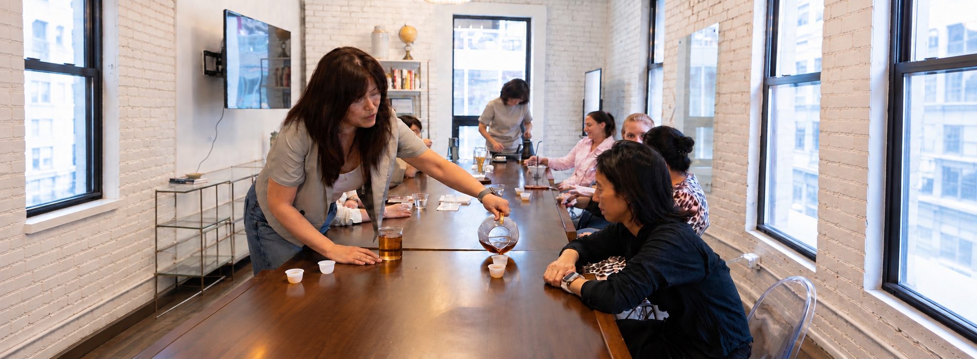 A host leans across a long wooden table, pouring tea into small cups for a group of guests in a bright loft with white brick walls.