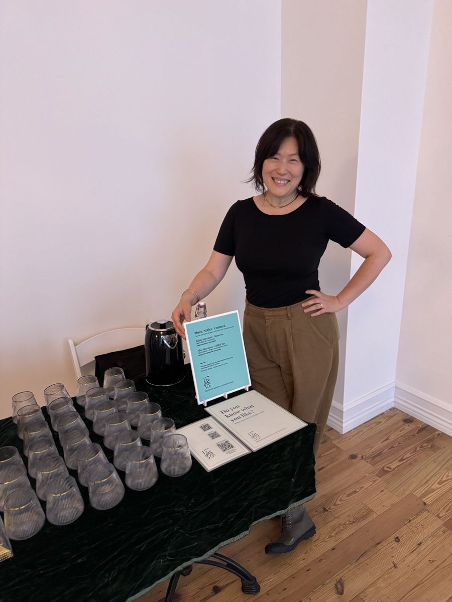 A smiling woman beside a velvet-covered table set with rows of glass cups, a kettle and 143 Labs signs reading “Move. Notice. Connect.” and “Do you know what you like?”