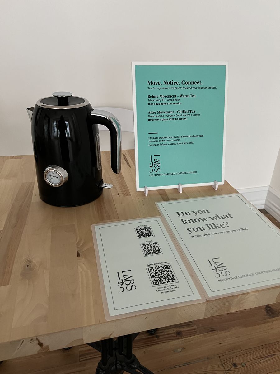 A black kettle on a wooden table beside 143 Labs signs: a turquoise card reading “Move. Notice. Connect.”, a QR-code card and one asking “Do you know what you like?”