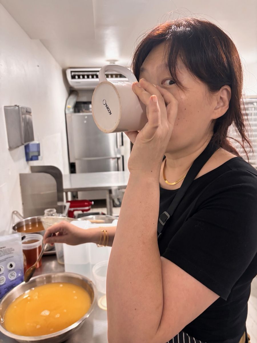 A woman in a black T-shirt and apron tastes from a white mug in a commercial kitchen, a bowl of orange liquid on the counter beside her.